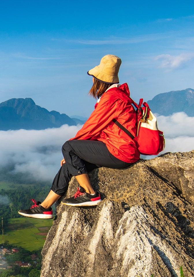 Person in a red jacket and beige hat sitting on a rocky cliff overlooking fog-covered mountains and a valley below.